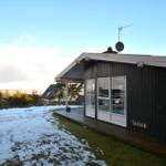 Schwarzes Holzhaus mit Terrasse und Schneebedecktem Grundstück. Satellitenantenne und Fenster sichtbar.
