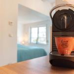 Kaffeemaschine mit Tasse auf Küchenarbeitsplatte. Im Hintergrund ist ein Schlafzimmer mit Bett und Fenster sichtbar.