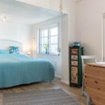 Schlafzimmer mit Bett, Schreibtisch und Fenstern. Holzboden und weiße Wände.