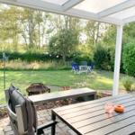 Terrasse mit Tisch, Stühlen und Grill unter Pavillon. Grünfläche mit Gartenmöbeln im Hintergrund.