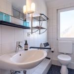 Badezimmer mit Waschbecken, Toilette und Fenster. Weiße Fliesen und blaue Accessoires.