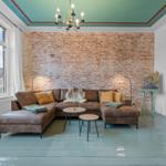 Ein Wohnzimmer mit braunem Sofa, Backsteinwand und grünem Boden. Decke mit Kronleuchter.