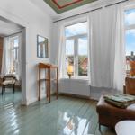 Wohnzimmer mit braunem Sofa und Blick auf die Straße. Holzfußboden und Fenster mit weißen Vorhängen.