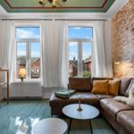 Wohnzimmer mit braunem Sofa, Holzfußboden und Backsteinwand. Große Fenster mit Blick auf Dächer.