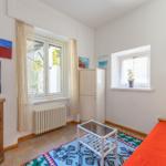 Zimmer mit Fenster, Schrank, TV und Bett mit roter Bettwäsche.