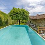Pool mit Liegestühlen und Tisch. Steinhaus mit Terrasse und grüner Umgebung.