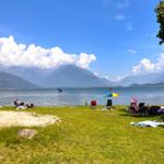Grüner Strand mit Liegestühlen und buntem Sonnenschirm am See. Im Hintergrund sind Berge und Wassersportler zu sehen.