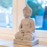 Eine Buddha-Statue sitzt auf einem Fensterbrett. Im Hintergrund ist ein Fenster mit Blick nach draußen.
