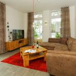 Wohnzimmer mit braunem Sofa, Holztisch und Fernseher. Große Fenster mit Blick auf Garten.