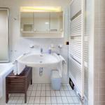 Badezimmer mit Badewanne, Waschbecken und Spiegel. Heizkörper und Holztür sind sichtbar.