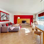 Wohn- und Essbereich mit Sofa, TV und Tisch. Rote Wand und Bergbilder.