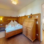Schlafzimmer mit Holzbett, Schrank und Nachttisch. Tür führt in einen Flur mit Spiegel.