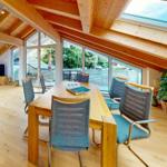 Ein Esszimmer mit Holzdecke, Tisch und Stühlen. Durch große Fenster ist eine Terrasse sichtbar.