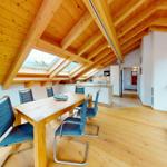 Dining room with wooden table, blue chairs, and kitchen in attic space with wooden beams and skylights.