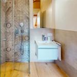 Dusche mit Glaswand, Waschbecken und Spiegel. Holzfußboden und Wandfliesen.