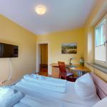 Zimmer mit Bett, Schreibtisch, Fernseher und Fenster mit Jalousien.