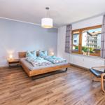 Schlafzimmer mit Doppelbett, Holzboden und Sessel. Fenster mit Vorhängen und Blick auf Gebäude.