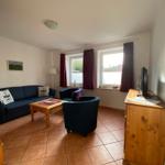 Wohnzimmer mit Sofa, zwei Sesseln, Holztisch und TV-Schrank. Fenster mit roten Vorhängen.