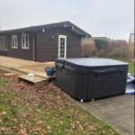 Schwarzer Whirlpool auf Terrasse neben Holzhaus mit grünem Rasen und herbstlichen Blättern.