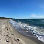Strand mit Wellen, Sand und klarem Himmel. Ein paar Menschen sind fern sichtbar.