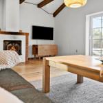 Wohnzimmer mit Kamin, TV und Holztisch. Große Fenster mit Blick auf Außenbereich.