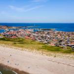 Strand mit Dünen und Küstenstadt mit Häusern und Hafen.