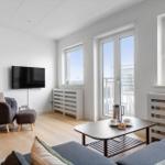 Wohnzimmer mit Sofa, Sessel, Couchtisch und Fernseher. Große Fenster und Balkontür.