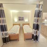 Zwei Einzelbetten mit Holzrahmen und gestreiften Vorhängen. Küche im Hintergrund sichtbar.