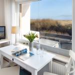 Wohnraum mit Tisch, Stühlen und TV. Durch Fenster Blick auf Strand und Dünen.