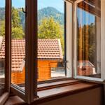 Durch das Fenster blickt man auf ein Holzhaus mit rotem Dach und grüne Berghänge.