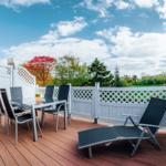 Terrasse mit Tisch, Stühlen und Liegestuhl. Weiße Brüstung, Blick auf Bäume und Himmel.