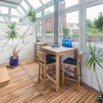 Ein Raum mit Holzboden, Tisch, Stühle und Pflanzen. Große Fenster bieten Blick auf Außenbereich.