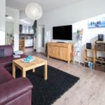 Wohnzimmer mit Sofa, Couchtisch und Fernseher. Küche im Hintergrund sichtbar.