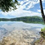 Stiller See mit Spiegelung des Himmels, umgeben von Bäumen und grüner Landschaft.