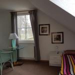 Schlafzimmer mit Dachschräge, Bett, Nachttisch, Stuhl und Tisch unter Fenster.