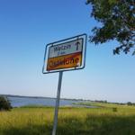 Schild weist auf Welzin und Ostklüne hin, mit Blick auf Wasser und Grasland.