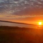 Sonnenuntergang über einem See mit bewölktem Himmel und Gras im Vordergrund.
