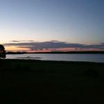 Sonnenuntergang über einem See mit Uferbereich und Bäumen im Vordergrund.