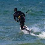 Ein Kitesurfer fährt auf dem Wasser mit einer weißen Board und einem schwarzen Wetsuit.