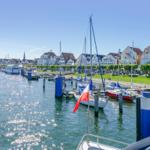 Hafen mit Segelbooten, grüner Uferweg und Häusern im Hintergrund.