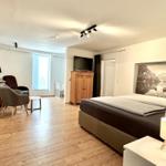 Schlafzimmer mit Bett, Fernseher und Wandbild. Holzboden und Sessel im Raum.