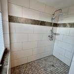 Dusche mit weißen Fliesen und dunkler Zierleiste. Bodenfliesen und Duschbrause sichtbar.