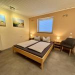 Schlafzimmer mit Doppelbett, zwei Nachttischen, Sessel und Fenster mit Jalousie.