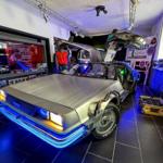 Ein silberner DeLorean mit geöffneten Türen steht in einem Raum mit blauer Beleuchtung.