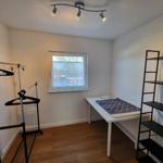 Kleines Zimmer mit Tisch, Kleiderständer und Regal. Fenster mit Vorhang. Holzboden.