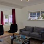 Wohnzimmer mit Sofa, zwei schwarzen Stühlen und Glas-Tisch. Fenster mit roten Vorhängen.