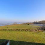 Green field with stone wall, view of sea and trees under blue sky.