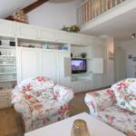 Living room with floral seating, white cabinets, and TV. Mezzanine with balustrade visible.