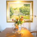 Dining table with flowers, bottles, and glasses in front of a garden-themed painting.