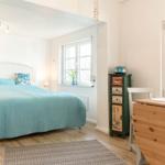 Schlafzimmer mit Bett, Schreibtisch und Fenstern. Holzboden und Wandfarbe weiß.